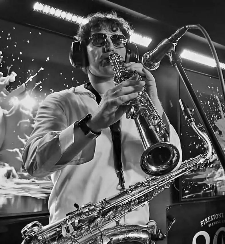 Black-and-white photo of the saxophonist playing soprano sax into a microphone on stage, with a tenor sax in front